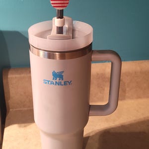 May include: A light pink Stanley tumbler with a handle and a clear lid. A straw is inserted through the lid, topped with a decorative acrylic topper shaped like a mouse head featuring a red, white, and blue American flag pattern.
