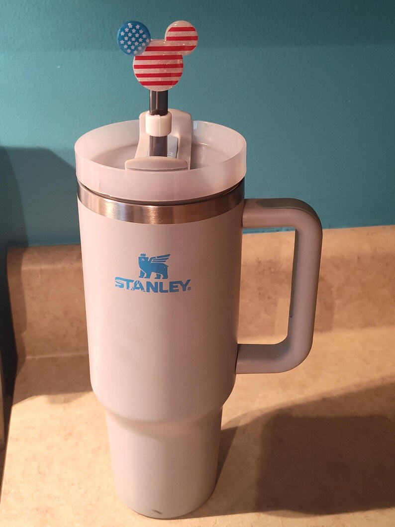 May include: A light pink Stanley tumbler with a handle and a clear lid. A straw is inserted through the lid, topped with a decorative acrylic topper shaped like a mouse head featuring a red, white, and blue American flag pattern.