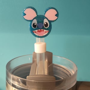 May include: A blue glittery Stitch-themed straw topper with pink ears and a wide grin, attached to a clear straw. The straw is inserted into a gray tumbler with a clear lid, set against a light blue background.