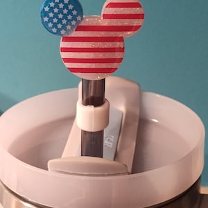 May include: A straw topper featuring a mouse-shaped head with a glittery finish. One ear is blue with white stars, while the other ear and the main head section feature red and white horizontal stripes. The topper is attached to a dark straw.