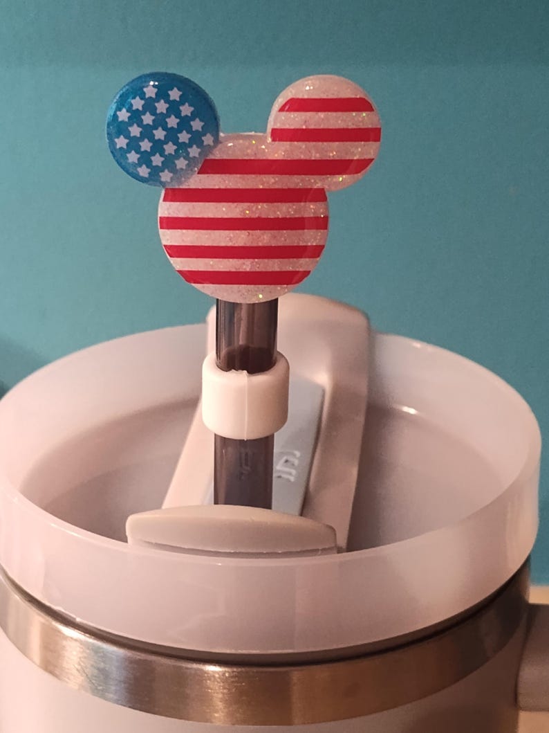 May include: A straw topper featuring a mouse-shaped head with a glittery finish. One ear is blue with white stars, while the other ear and the main head section feature red and white horizontal stripes. The topper is attached to a dark straw.
