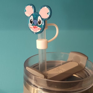 May include: A blue glittery Stitch-themed straw topper with pink ears and a white straw. The topper is attached to a gray tumbler with a clear lid. The background is a solid teal color.