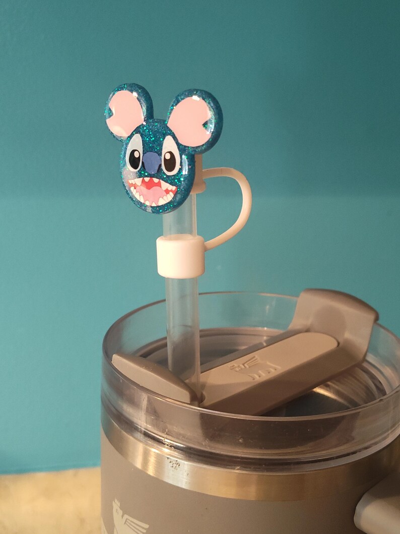 May include: A blue glittery Stitch-themed straw topper with pink ears and a white straw. The topper is attached to a gray tumbler with a clear lid. The background is a solid teal color.