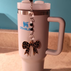 May include: A light gray Stanley tumbler with a handle and a clear straw. A charm with clear and black beads and a leopard print bow is attached to the lid. The Stanley logo is visible on the front.