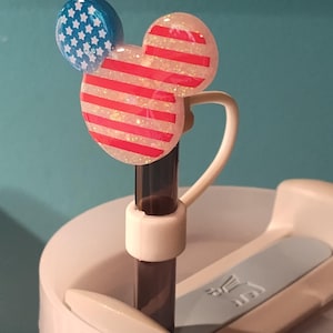 May include: A straw topper shaped like a mouse head with a glittery finish. One ear is blue with white stars, and the main head features red and white horizontal stripes. The topper is attached to a dark straw inserted into a white tumbler lid.