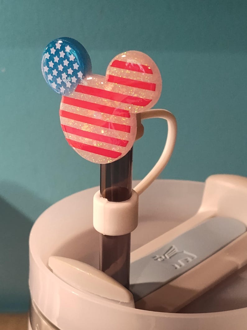 May include: A straw topper shaped like a mouse head with a glittery finish. One ear is blue with white stars, and the main head features red and white horizontal stripes. The topper is attached to a dark straw inserted into a white tumbler lid.