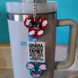 Puede incluir: Un vaso blanco con tapa y asa negras. Un charm con una cabeza de Mickey Mouse roja y blanca, una cabeza de Stitch azul y una etiqueta blanca con el texto "Ohana Means Family Family Means Nobody Gets Left Behind Or Forgotten" est&aacute; unido a la tapa.