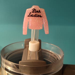 May include: A pink glittery jacket-shaped drink charm with the words "Pink Ladies" in black script. The charm is attached to a clear straw and sits atop a stainless steel tumbler with a clear lid. The background is a solid teal color.