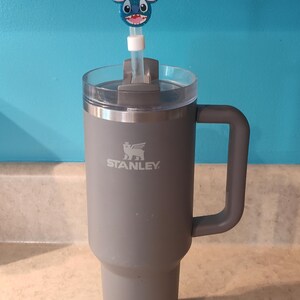 May include: A gray Stanley tumbler with a handle and a clear lid. A blue and pink character straw topper is inserted into the lid. The tumbler is on a light-colored countertop with a blue wall in the background.