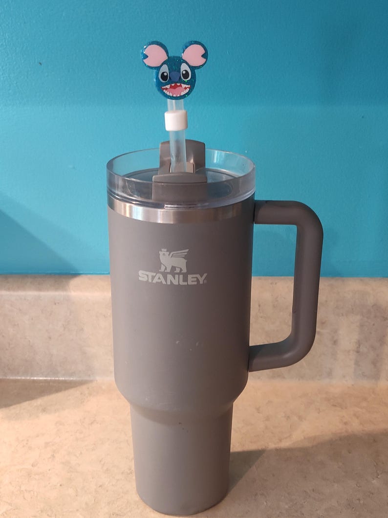 May include: A gray Stanley tumbler with a handle and a clear lid. A blue and pink character straw topper is inserted into the lid. The tumbler is on a light-colored countertop with a blue wall in the background.
