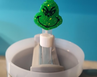 May include: A green Grinch-themed drink charm with glitter accents, featuring a smiling face and a mischievous expression. The charm is attached to a clear straw, placed inside a light blue and silver travel mug.
