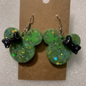 May include: A pair of green glitter earrings in a mouse ear shape, with a black bow and a green frog silhouette. The earrings hang from silver-colored fish hook ear wires. The earrings are displayed on a brown card.