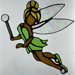 Puede incluir: Un hada de vidrio emplomado con alas verdes y un cuerpo marrón que sostiene una varita con una esfera blanca. El hada tiene el pelo amarillo y está volando.