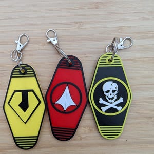 May include: Three keychains with silver clasps. One is yellow with a black diamond and a black letter J. The second is red with a white design. The third is black and yellow with a skull and crossbones.