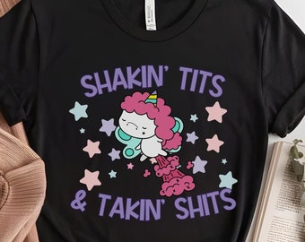 May include: Black t-shirt with the text "Shakin' Tits & Takin' Shits" and a cartoon unicorn graphic. The unicorn has a pink mane and tail, and is surrounded by stars. A mug and a sweater are next to the shirt.