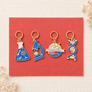 May include: Four science-themed keychains with gold-colored rings. The charms are blue, white, and yellow, featuring a flask, microscope, globe with "-CHEMISTRY-" banner, and DNA strand. The keychains are on a red background.
