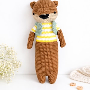 May include: A handmade crochet otter doll with a brown body, head, and arms. The otter wears a yellow and white striped shirt and a light green vest. The doll has a black nose and a smiling mouth. The background is white.