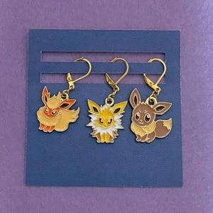 May include: Three gold-colored earrings featuring cartoon characters. The earrings are in the shape of a fox, a yellow creature with a spiky collar, and a brown and tan creature. The earrings are displayed on a blue card.