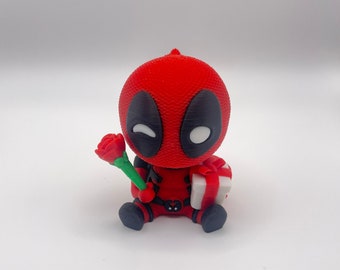 May include: A red and black Deadpool figurine holding a red rose and a white gift box. The figurine is winking and has a mischievous expression.