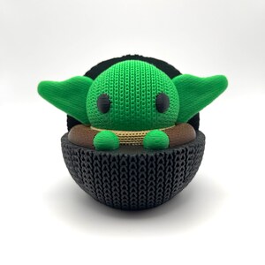 Peut inclure: Une figurine imprimée en 3D verte et noire d'un personnage de la franchise Star Wars. Le personnage est assis dans une capsule noire avec un bord brun.