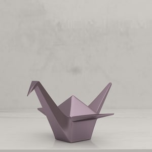 May include: A lavender origami crane sculpture. The paper bird is folded with sharp angles and a pointed beak. The crane is displayed on a white surface against a neutral background. This decorative item is a symbol of peace and longevity.