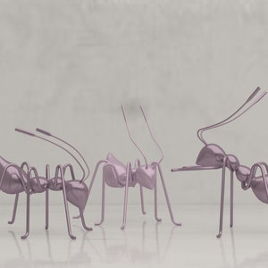 Puede incluir: Tres hormigas rosadas metálicas hechas de alambre, de pie sobre una superficie blanca. Las hormigas están mirando en diferentes direcciones y tienen patas largas y delgadas.