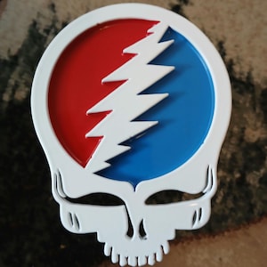 May include: White metal belt buckle with a red, white, and blue lightning bolt design. The lightning bolt is inside a circle and the buckle is shaped like a skull.