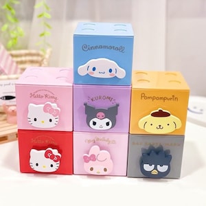 May include: Six colorful stackable storage boxes featuring different Sanrio characters. The boxes are pink, red, purple, blue, orange, and gray. Each box has a different character on the front, including Hello Kitty, Cinnamoroll, Kuromi, My Melody, Pompompurin, and Badtz-Maru.