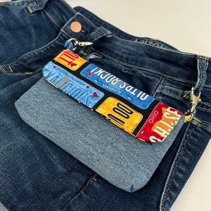 May include: A small denim pouch with a colorful fabric lining featuring a pattern of vintage license plates from different states. The pouch is attached to a pair of blue jeans with a metal ring and a key chain.