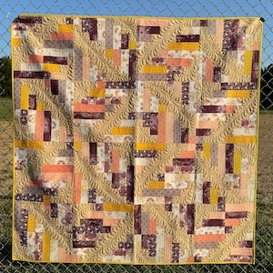 May include: A handmade quilt with a patchwork design. The quilt features rectangular blocks in shades of yellow, peach, burgundy, and cream. The blocks are separated by a tan fabric with a leaf pattern. The quilt is bordered with a yellow binding.