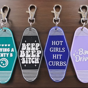 May include: Set of five keychains with different sayings. The keychains are shaped like motel room keys. The sayings are "Having a Menty B", "Beep Beep Bitch", "Hot Girls Hit Curbs", "Bad Driver", and "I'm Not a Ho, I'm a Goddess".
