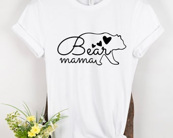 May include: White t-shirt with a black graphic of a bear silhouette and the words "Bear mama". The bear has three heart-shaped accents. The shirt is hanging on a wooden hanger.