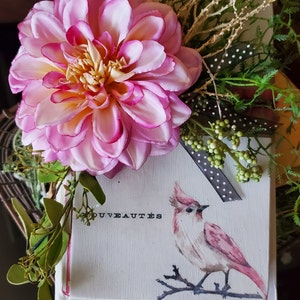 Puede incluir: Una caja de madera blanca decorada con una flor rosa, hojas verdes y una cinta de lunares negros y blancos. La caja tiene una ilustración de un pájaro rosa y el texto "Nouveautés" impreso en ella.
