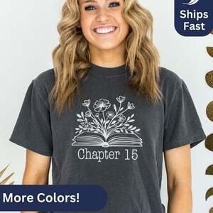 May include: A dark gray t-shirt with a white graphic of an open book with flowers growing out of it. The text "Chapter 15" is printed below the book.