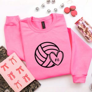 May include: A bright pink sweatshirt with a black volleyball graphic and a heart with the number 13. The sweatshirt is surrounded by decorative items, including a pink notebook, macarons, and a tumbler.