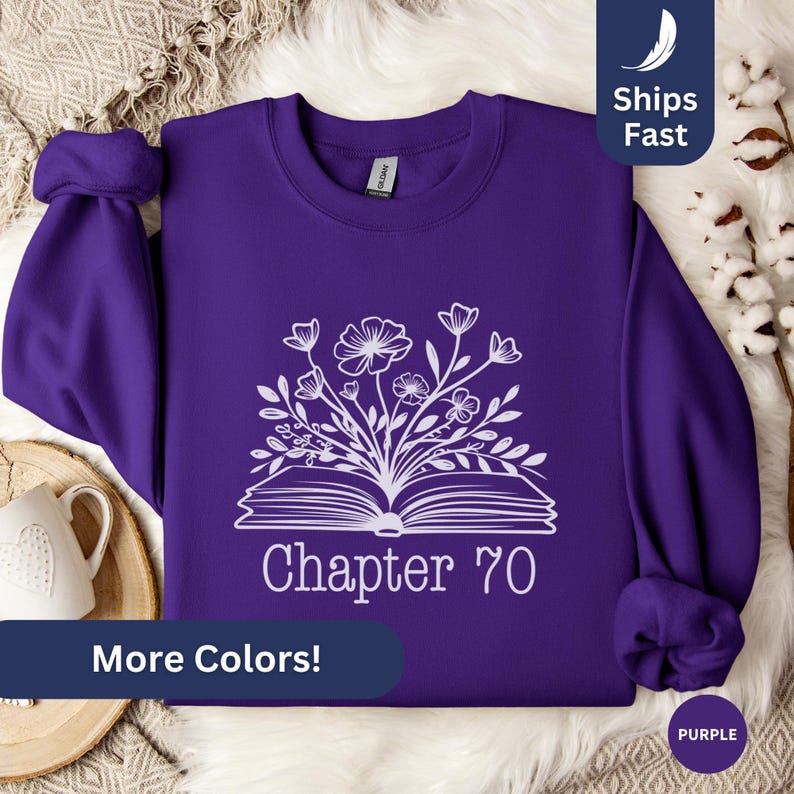 May include: Purple sweatshirt with a white design of an open book with flowers growing out of it. The text "Chapter 70" is below the book. The sweatshirt is on a white fuzzy surface.