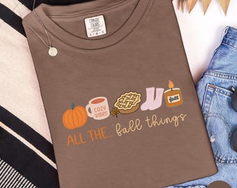 May include: Espresso-colored t-shirt with a graphic of fall-themed icons: a pumpkin, mug with "Cozy Vibes", pie, socks, and a candle. The text "ALL THE... fall things" is printed below the icons. The shirt is a Comfort Colors brand.