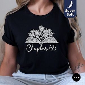May include: Black t-shirt with a white design of an open book with flowers growing out of it. The text "Chapter 65" is below the book.