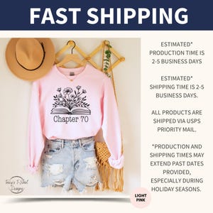 May include: A light pink sweatshirt with a black graphic of an open book with flowers growing out of it. The text "Chapter 70" is printed below the book. The sweatshirt is hanging on a wooden rack with a brown hat and a woven bag.