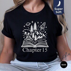 May include: Black t-shirt with a white graphic of a bear, boots, trees, mountains, a rocket, and a planet above an open book. The text "Chapter 15" is below the book. The shirt has a "Super Soft" label.