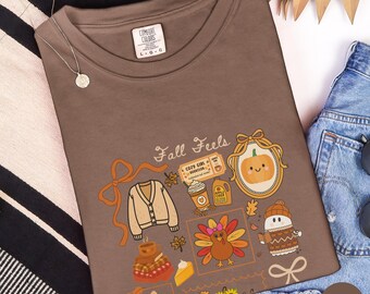 May include: Espresso-colored Comfort Colors t-shirt with the words "Fall Feels" and autumn-themed illustrations. The design includes a pumpkin, turkey, sweater, and other fall elements. The shirt is size L-G-G.