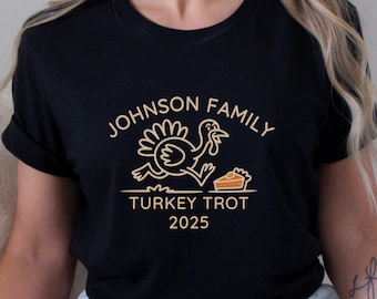 May include: Black t-shirt with a gold graphic. The graphic includes the text "JOHNSON FAMILY TURKEY TROT 2025" with a cartoon turkey running towards a slice of pie. The shirt is a crew neck.