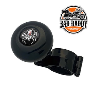 May include: Black steering wheel knob with a silver spider web design and a red hourglass in the center. The knob is attached to a black clamp. The text "BAD DADDY GARAGE" is visible in the background.