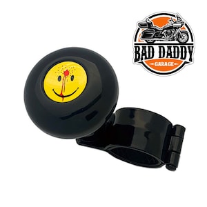 May include: Black steering wheel knob with a yellow smiley face with a red blood splatter on the forehead. The knob has a black metal clamp.