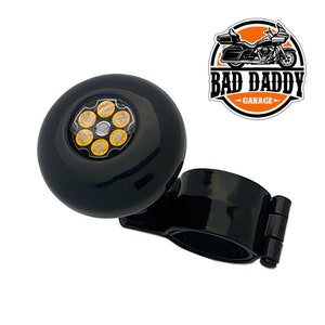 May include: Black motorcycle handlebar grip with a circular design featuring six gold-colored bullet casings. The grip is mounted on a black bracket. The Bad Daddy Garage logo is in the upper right corner.