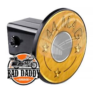 May include: A gold-colored metal hitch cover with a black rim. The cover features a stylized image of a .44 Magnum bullet casing with the text "44 MAG" and three gold stars. The cover is attached to a black hitch receiver.