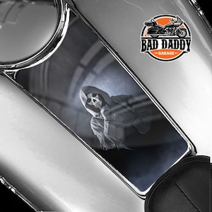 May include: A gas tank decal featuring a hooded figure with a skull face, holding a finger to its lips. The decal has a dark, smoky background and the text "Bad Daddy Garage" in a circle with a motorcycle graphic.