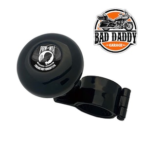 May include: Black steering wheel knob with a POW MIA logo and the text "You are not forgotten". The knob is attached to a black clamp. The logo is a white silhouette of a soldier's head with a barbed wire fence and the words POW MIA.