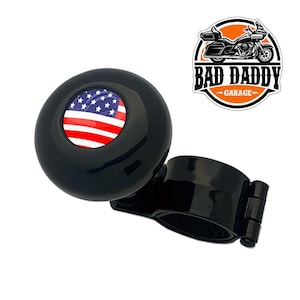 May include: Black steering wheel knob with a red, white, and blue American flag design. The knob is attached to a black clamp.