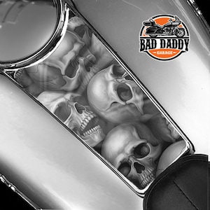 May include: A black and white graphic of a motorcycle gas tank with a skull design. The design features multiple skulls in a pile, with the text "Bad Daddy Garage" in a circle above the skulls.